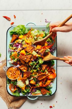 two hands holding wooden spoons over a plate of food with lettuce, carrots and meat