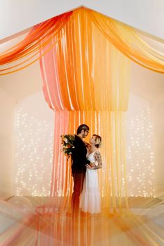 a bride and groom standing under an orange drape