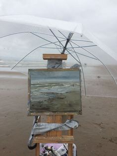 an easel is set up on the beach to display some paintings and umbrellas