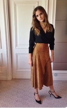 a woman standing in front of a door wearing a black shirt and brown skirt with high heels