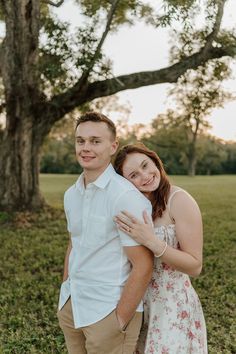 a man and woman standing next to each other in the grass under a large tree