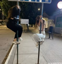 two women sitting on top of poles near a street sign at night, with one leaning over the pole