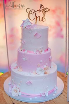 a three tiered cake with pink and white flowers on the bottom is displayed in front of a one sign