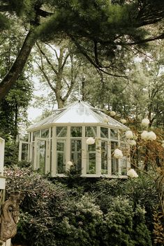 a glass house surrounded by greenery and trees in the woods, with white flowers growing around it