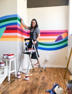 a woman standing on a ladder painting a wall with colorful stripes and rainbows in the background