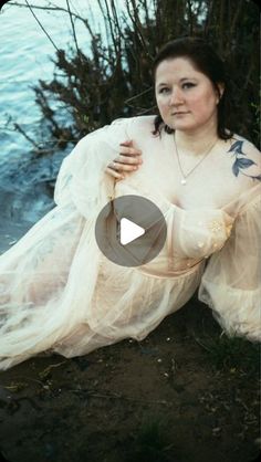 a woman in a white dress sitting on the ground next to some bushes and water