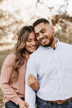 a man and woman smile at the camera while standing in front of trees with their arms around each other