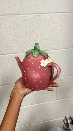 a hand holding a ceramic teapot with flowers painted on the top and bottom, in front of a white brick wall