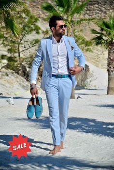 a man in a blue suit walking down the beach with his shoes on and sunglasses on