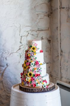 a wedding cake with flowers on it sitting on top of a white barrel next to a brick wall