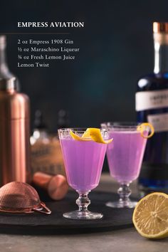 two glasses filled with purple lemonade next to a bottle and copper flasks