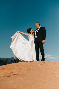 a bride and groom standing on top of a rock in the desert with their arms around each other