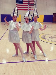 three girls in silver dresses and hats on a basketball court with an american flag behind them