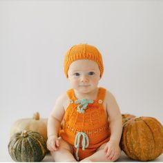 a baby wearing an orange knitted romper and matching hat sits in front of pumpkins