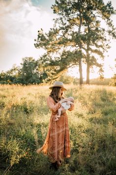 a woman in a dress and hat holding a baby while standing in a field with trees