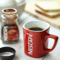 a red coffee mug sitting on top of a table next to a jar of sugar