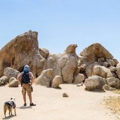 a man standing next to a dog on top of a dirt field near large rocks