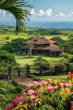 a large house surrounded by lush green trees and flowers in the foreground, with an ocean view behind it