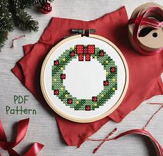 a cross stitch christmas wreath on a red cloth next to some ribbon and a spool of thread