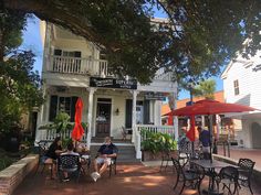 people sitting at tables in front of a white house with red umbrellas on the porch