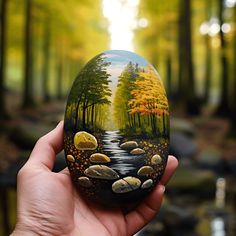 a hand holding an easter egg painted with trees and rocks in the forest, on a sunny day