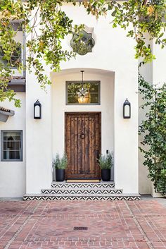 a white house with a wooden door surrounded by greenery and potted plants on the front porch