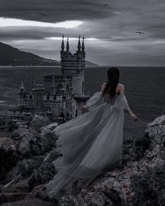 a woman standing on top of a rocky cliff next to the ocean with a castle in the background