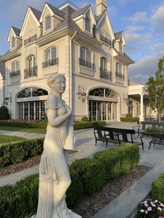 a statue in front of a large white house with lots of bushes and flowers around it