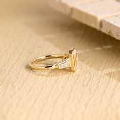 a yellow gold engagement ring with an emerald cut diamond on the side, sitting on a piece of wood