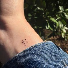 a small cross tattoo on the foot of a person's left ankle in front of some bushes