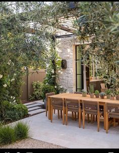 an outdoor dining table surrounded by greenery and trees in front of a stone building