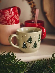 two mugs with trees painted on them are sitting next to pine cones and evergreen branches