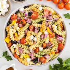 a bowl filled with pasta salad next to tomatoes, olives and garlic on a white surface