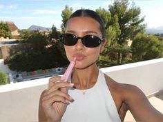 a woman in sunglasses brushing her teeth with a pink toothbrush on the outside balcony