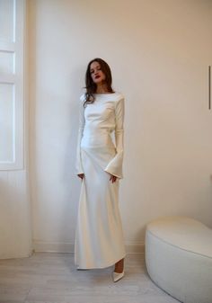 a woman standing in front of a white wall wearing a long white dress and heels