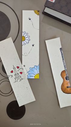 three bookmarks with drawings on them sitting on a table next to an open book