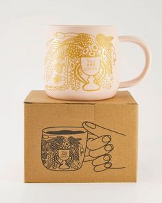 a pink coffee mug sitting on top of a cardboard box next to a golden cup