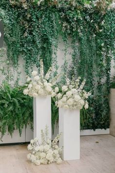 two white vases with flowers and greenery in front of a wall that has ivy growing on it