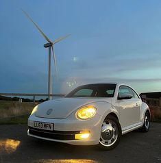 a white volkswagen beetle parked in front of a wind turbine at dusk with the headlights on