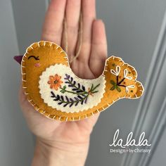 a hand holding an embroidered bird ornament in it's left hand, with flowers and leaves on the wings