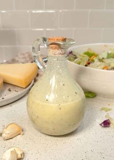 a glass bottle filled with dressing next to a bowl of salad and cheese on a counter