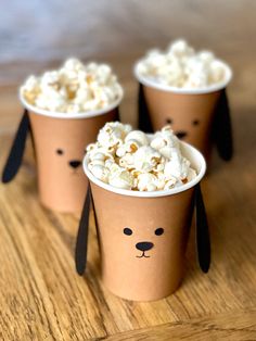 three paper cups with popcorn in them on a wooden table, one is shaped like a dog and the other is made to look like a bear