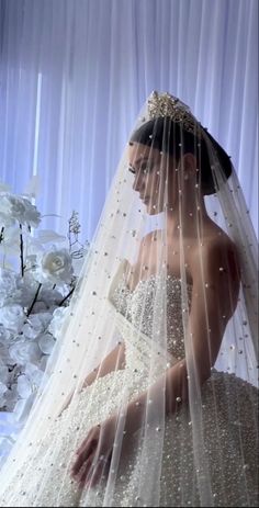 a woman wearing a wedding dress and veil in front of white flowers with sheer curtains behind her