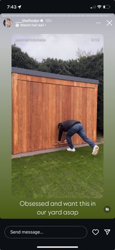 a man leaning over to pick up something from the grass in front of a wooden shed