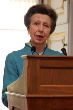 an older woman standing at a podium with microphones in front of her and looking off into the distance