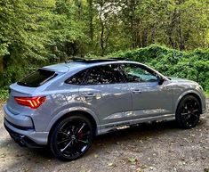 the rear end of a silver car parked in front of trees and bushes on a dirt road