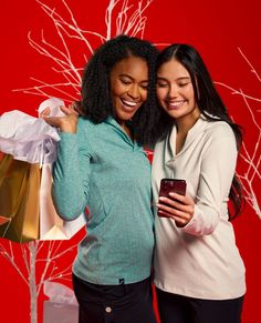 two women standing next to each other holding shopping bags and looking at a cell phone