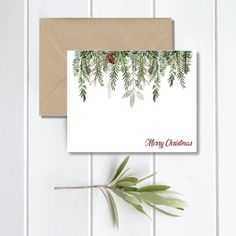 a christmas card with evergreen branches and pine cones on it, next to a brown envelope