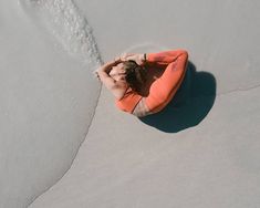 a woman in an orange shirt is sitting on the beach with her hands behind her head