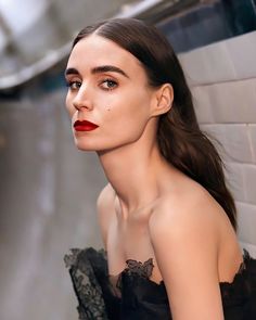 a woman with long hair wearing a black dress and red lipstick, leaning against a white brick wall